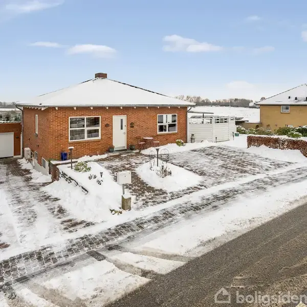 En rød murstensvilla står stille i et snedækket nabolag med en parkeringsplads foran og en garage til venstre, omgivet af andre huse og en vej.