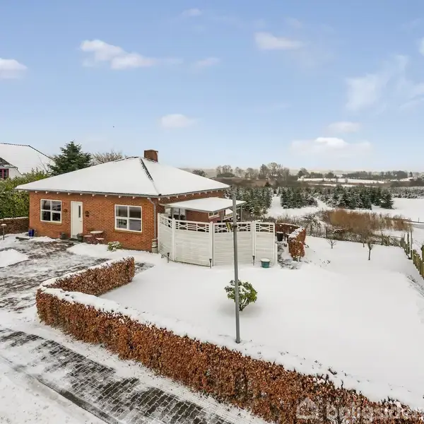 Et rødt murstenshus står omgivet af en snedækket have og terrasse. Hækken langs indkørslen er delvist snedækket. I baggrunden ses et åbent, snedækket landskab med få huse.