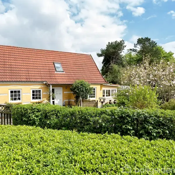 Gult hus med rødt tag står i solskin; omgivet af frodige buske og grønne træer. En have med blomstrende planter ses foran, mens himlen er let overskyet med blå sektioner.
