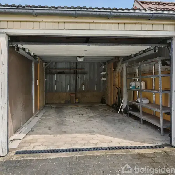 En åben garage med hylder fyldt med flasker og værktøj til højre. Beliggende i en villaindkørsel med en flisebelagt gulv og en træstruktur i baggrunden.