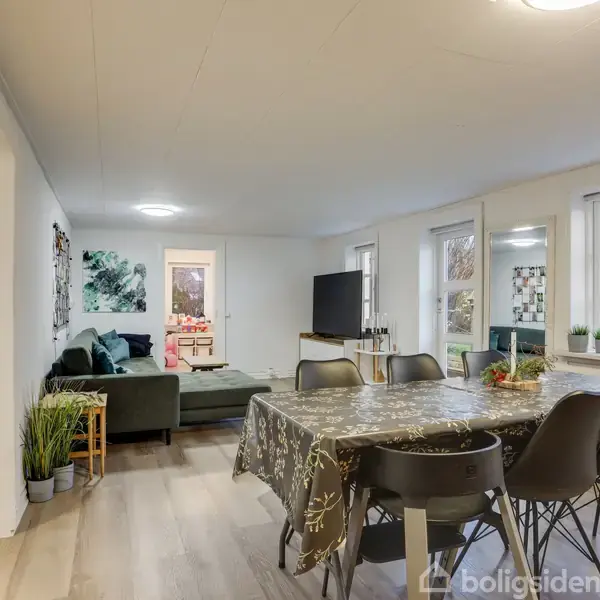 Spisebord med otte stole står i en lys stue med hvide vægge og store vinduer. En sofa og et fjernsyn ses i baggrunden. Flere planter pynter rummet.