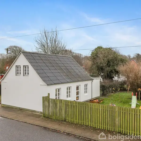 Et hvidt hus med sort tag står stille ved en vej. En græsplæne med legetøj og et lille hegn omkranser haven, omgivet af træer under en blå himmel.
