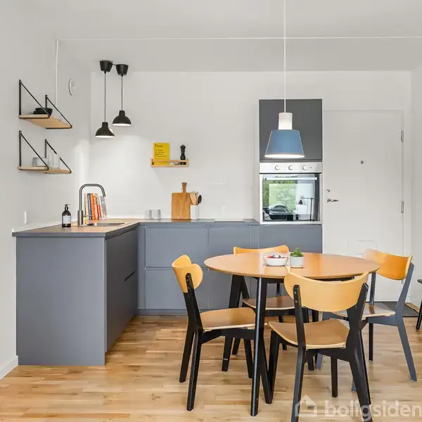 Et moderne køkken med et rundt spisebord og fire stole. Køkkenskabe er grå, med en indbygget ovn. Hylder og lamper på væggen. Plante og reol ved siden.