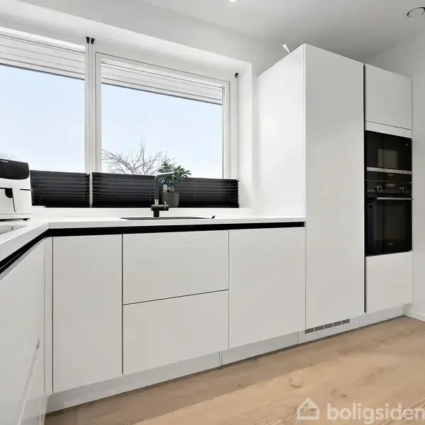 Et moderne, hvidt køkken indeholder en kaffemaskine og vask, hvor vaskemaskine er synlig i rummet ved siden af. Store vinduer lader naturligt lys trænge ind.