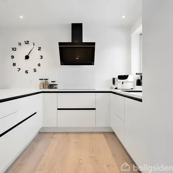 Et moderne køkken med hvide skabe. En stor urskive med tal er på væggen over en sort emhætte. Der står en kaffemaskine på bordet ved vinduet.