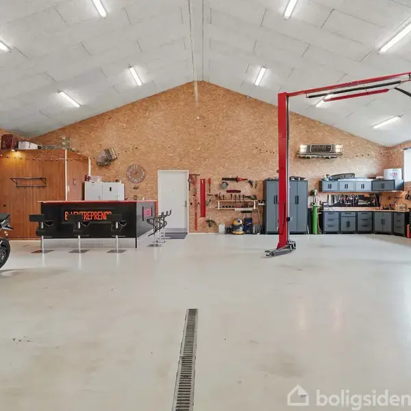 En stor garage med motorcykler til venstre og en bil-lift til højre. I baggrunden er der værktøj og opbevaringskasser langs væggene. Loftet har indbygget belysning.