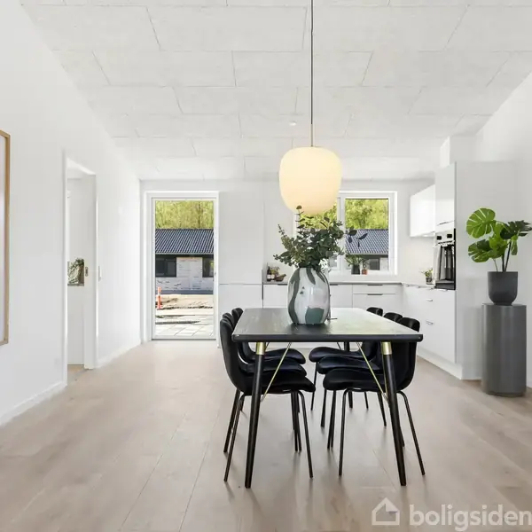 Et moderne spisestuemiljø med et sort spisebord og seks stole. En vase med grønne grene står på bordet. En stor lampe hænger over. Lysindfald fra vinduer. Plakat med teksten "KUBE".