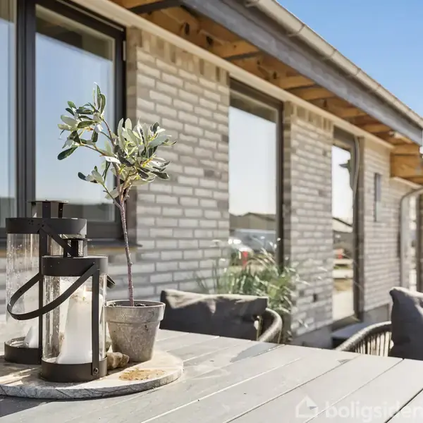 Lanterne står på et bord med en lille potteplante. Baggrunden viser en murstensbygning med flere vinduer, der reflekterer sollyset. Det ligner en terrasse ved et hus.