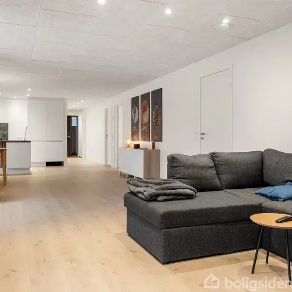 En sort sofa med puder står i en lys stue, hvor den vender mod et lille bord med en fjernbetjening. I baggrunden ses et spisebord og et åbent køkken.