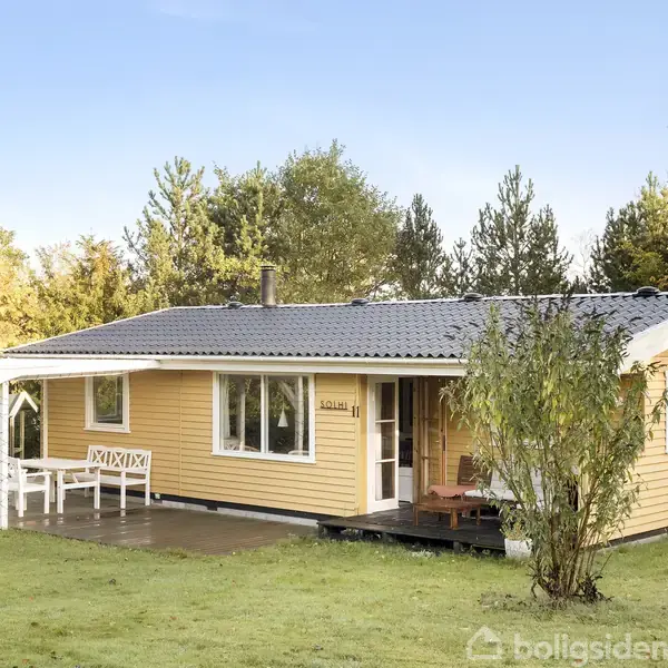 Et gult træhus med veranda står i en frodig have omgivet af træer. Et bord og bænk er placeret under verandaen. Skyfri blå himmel.