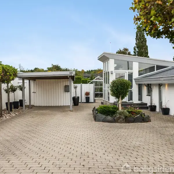Et moderne hus med store glaspartier står i en velholdt indkørsel, flankeret af potteplanter og små træer. Garage og skur til venstre, blå himmel i baggrunden.