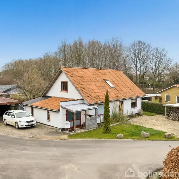 Hvid hus med rødt tag står stille ved en vej, omgivet af træer og andre huse. En hvid bil er parkeret i en carport til venstre.