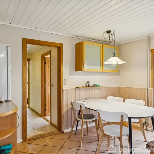 Et rundt spisebord med fire stole står i et lyst køkkenområde. En lampe hænger over bordet. Vinduet ved siden af viser en grøn have udenfor.