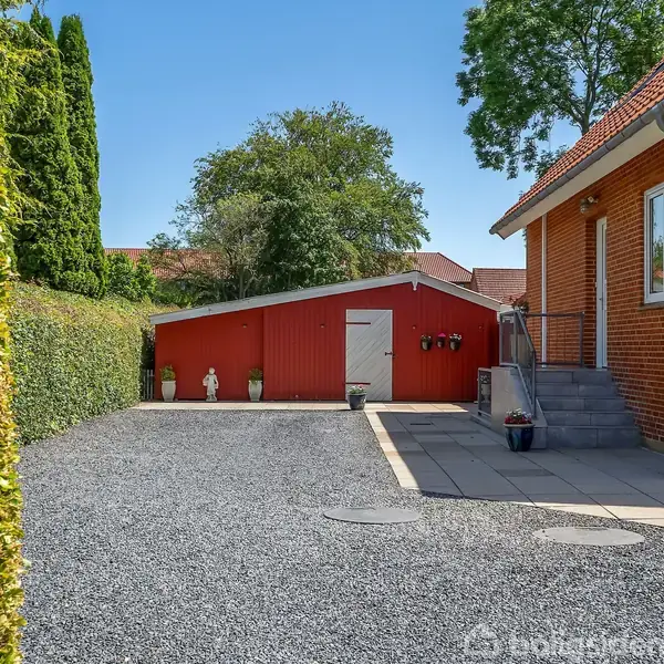 En rød murstensbygning ved siden af en grusbelagt indkørsel med en rød trægarage i baggrunden. Omkranset af grønne hække og træer, og der er blomsterdekorationer på trappen.