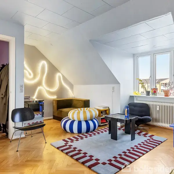Stue med moderne indretning. To sofaer står mod væggen med lysinstallation, bord i midten på tæppe med rødt mønster. Vindue til højre, yderligere to stole, reol og radiator.