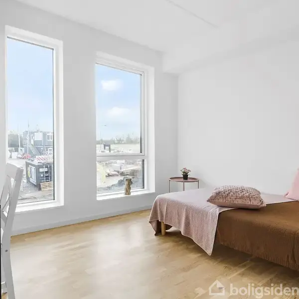 Et enkelt møbleret værelse med en daybed dækket af puder, står ved en væg. To vinduer giver lys og udsigt over byen. Et lille bord står ved siden af.