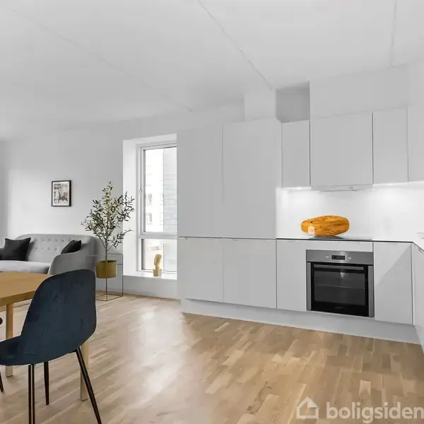 En moderne køkken-stue med hvide skabe og indbygget ovn. Et rundt spisebord med mørkeblå stole står til venstre. Sofa, planter og kunstværk findes i baggrunden ved vinduerne.