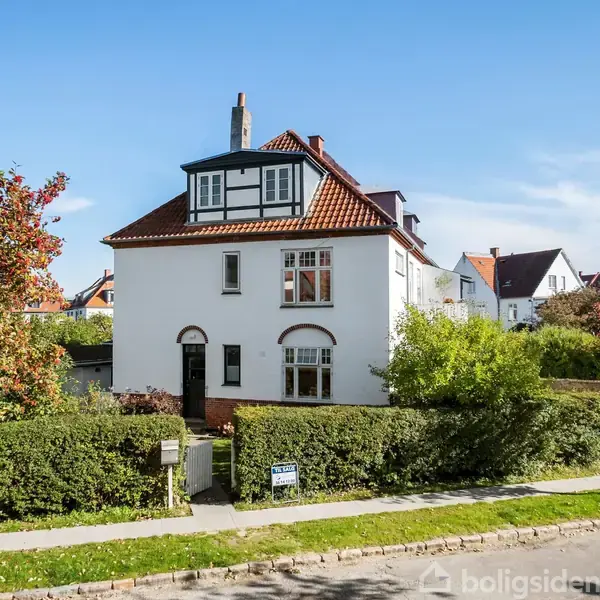 Hvidt to-etagers hus med rødbrunt tag, omgivet af grønne buske og træer. Et skilt foran informerer om "Til salg" og kontaktoplysninger for ejendomsmægleren. Blå himmel i baggrunden.
