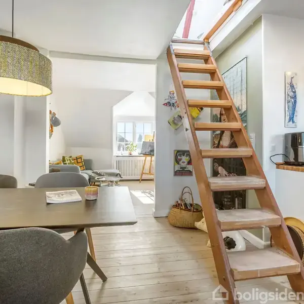 En trætrappe fører opad fra en hyggelig stue med spisebord, omgivet af stole og en lampe. Et blomstret arrangement står i en vase. Køkkenet ses i baggrunden.