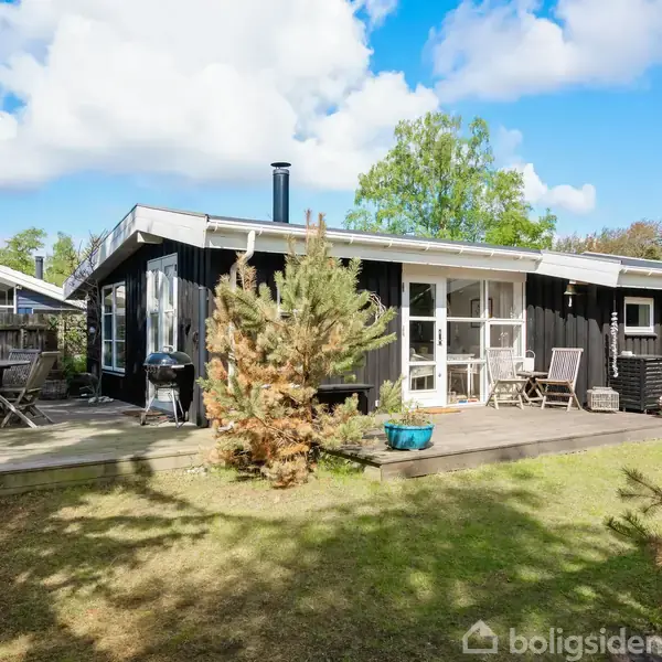 Sort træhus med terrasse, hvor der står et spisebord og stole. Haven er grøn med enkelte træer og buske omkring huset. Det er solrigt med blå himmel og skyer.