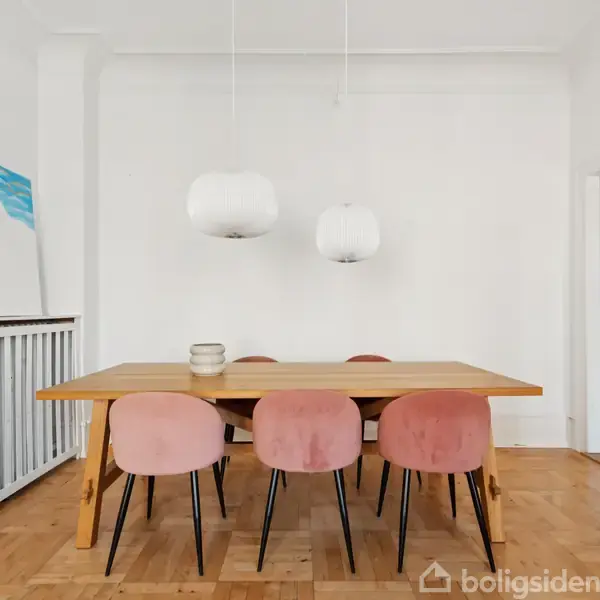 Et rektangulært træbord med seks lyserøde stole står i et lyst rum. To hvide lamper hænger over bordet. Et maleri med blå og hvide farver læner sig op ad væggen.