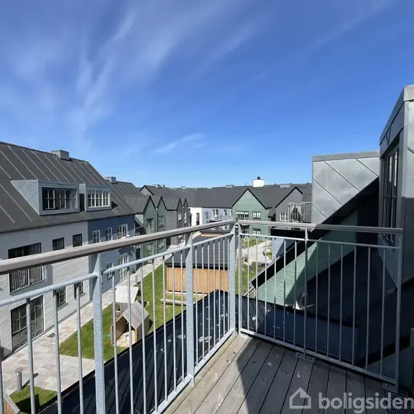 Altan med gitterværn, stående stille, tilbyder udsigt over moderne rækkehuse med grå tage og græsplæner i et solrigt boligområde.