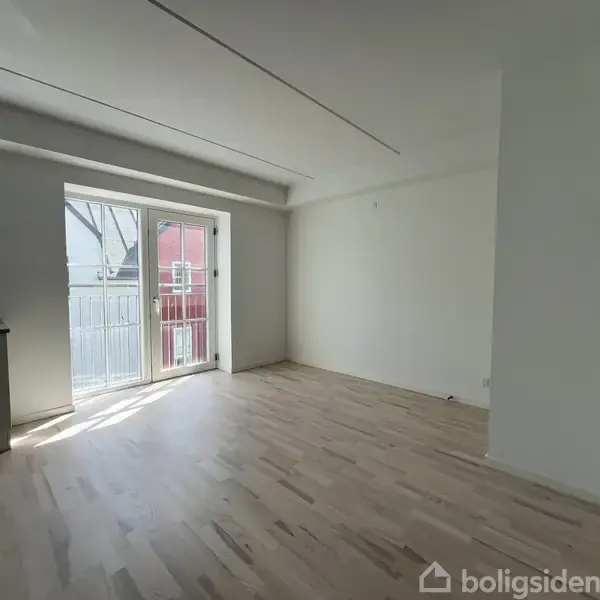 Et lyst, tomt rum med trægulv og store vinduer, der åbner ud mod en balkon. Væggene er hvide, og der er et åbent køkkenområde til venstre.
