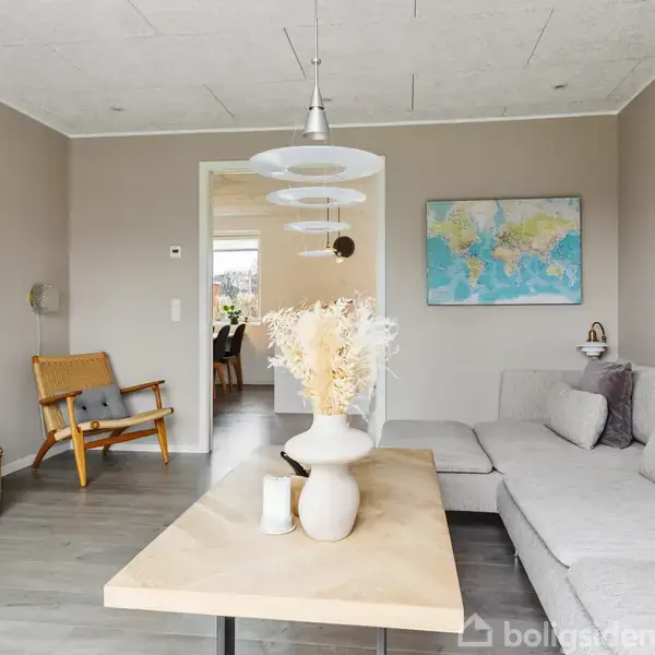 Gråt sofaarrangement med sofabord dekoreret med vase og lys. Væghængt fjernsyn til venstre. Kort på væggen bagved. Lys pendel hænger fra loftet. Trægulv og lænestol.