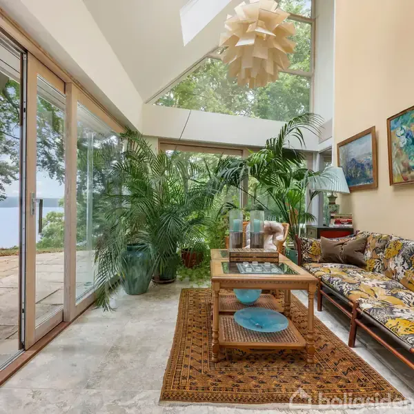 En stue med en sofa dekoreret med farverige puder står ved siden af et sofabord. Store glasdøre åbner til en terrasse med udsigt til skov og vand. Plante dekorerer rummet.