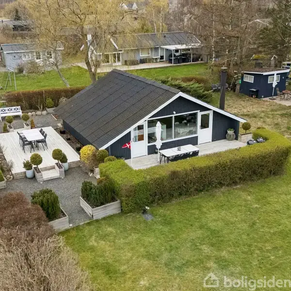 Sort-hus med skråtag står midt i have omgivet af hække. Terrassen har havemøbler og planter. Flere huse og træer ses i baggrunden, hvilket giver en fornemmelse af nabolag.