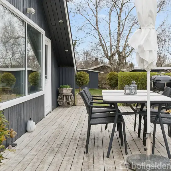 Terrasse med træbord og stole, omgivet af grønne buske. En parasol står ved bordet, og en grill er placeret til højre. Huset har mørk træbeklædning og store vinduer.