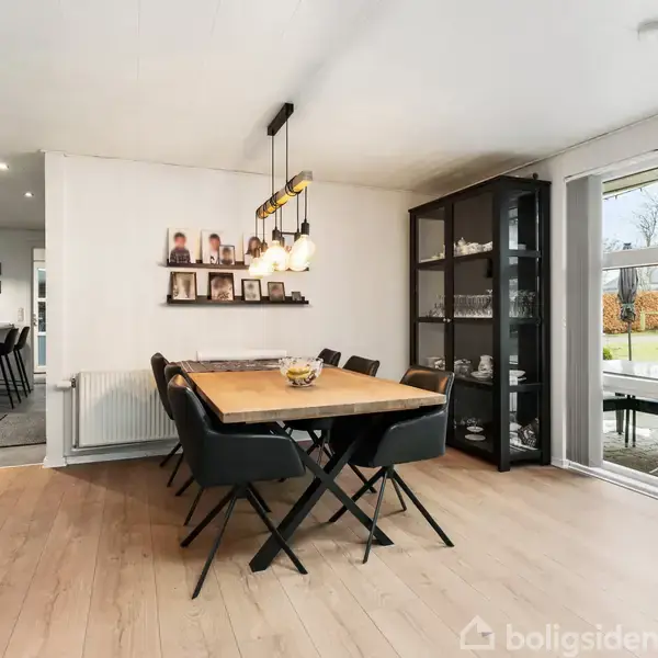 Spisebord med seks stole står i lys spisestue. Bordet har en skål i midten, og der er hylder med billeder på væggen. Glasmontre står til højre.