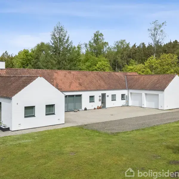 En hvid, moderne villa med rødt tegltag står på en stor, grøn græsplæne. Bygningen har store vinduer og en flisebelagt gårdsplads, omgivet af tæt, grøn skov.