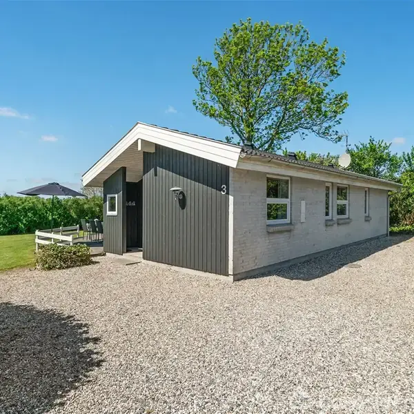 Et enkelt sommerhus står på en grusbelagt indkørsel, omgivet af grønne træer og buske. Huset har hvide vægge og sort træbeklædning omkring dørene. Nummeret 3 ses på væggen.