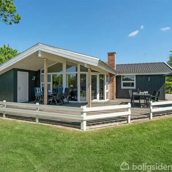 Sommerhus med træbeklædning står på grøn græsplæne. En terrasse har havemøbler og parasol. Omkring er der frodig vegetation og blå himmel.