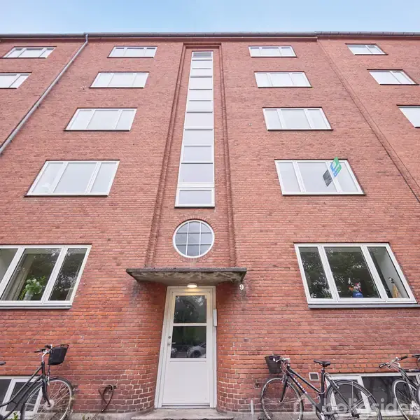 En rødstensbygning ses med flere vinduer, hvor cykler står parkeret foran indgangen. Bygningen har en karakteristisk høj vinduesrække i midten og omgives af en klar, blå himmel.