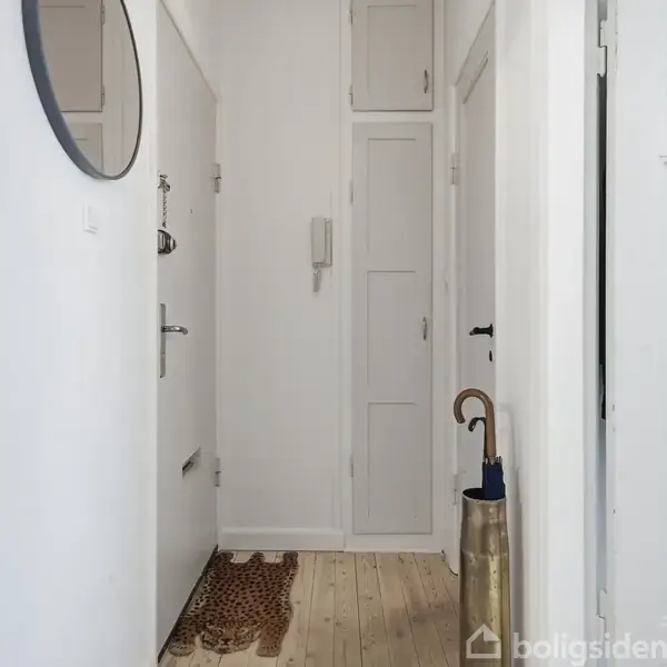 En smal entré med en rund spejl, en dørmåtte på trægulvet, og en paraply i en stander ved siden af en hvid væg. Væggene er lyse og minimalistiske.