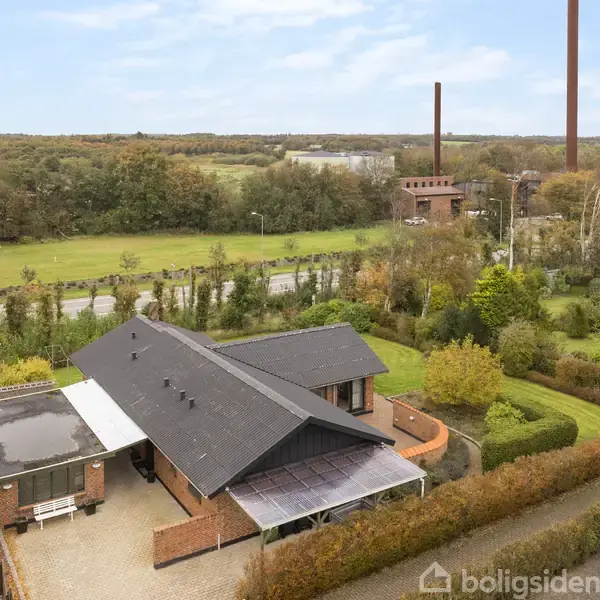 En murstensvilla med sort tag ligger omgivet af en velplejet have og hække. I baggrunden ses åbne marker, skov og industrielt prægede skorstene, der skaber en landlig atmosfære.