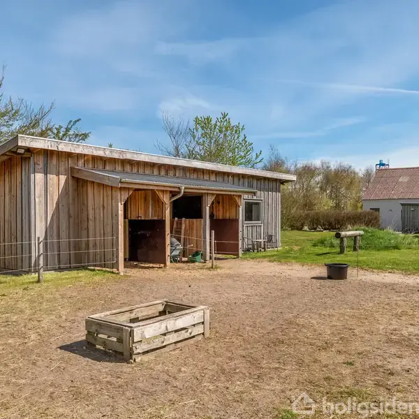 En træbygning med åbne staldområde står på en gårdsplads. Omkranset af hegn og græs, med en enkelt træramme og spand i forgrunden, omgivet af landskab og træer.