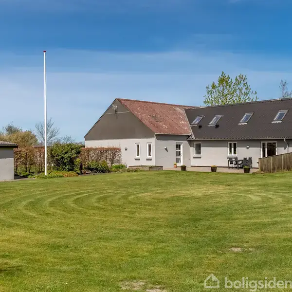 Et hvidt hus med sort tag står i midten af en velplejet græsplæne. Terrassen har havemøbler, omkranset af træer og en blå himmel.