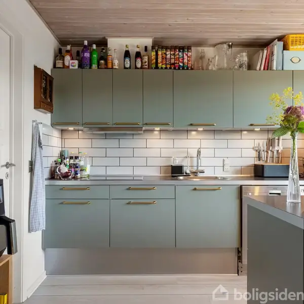 Køkken med grå skabe og hvid flisesplashback. Diverse krydderier og flasker står på hylderne. En frugtskål og en vase med blomster står på køkkenbordet, sammen med en kaffemaskine.