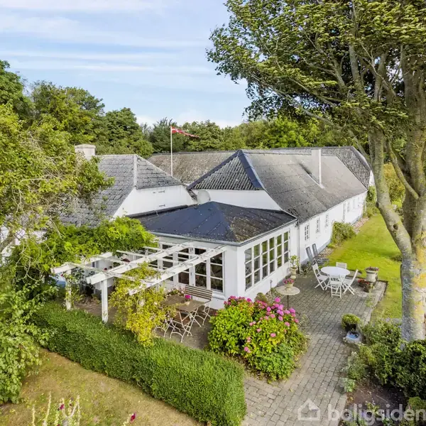 Hus med hvid facade og stråtag, placeret i en frodig have med havemøbler spredt på terrassen. Omgivet af høje træer og grønne marker i baggrunden.
