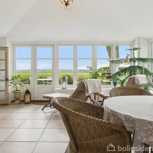 Et lyst rum med rattanmøbler og rundt bord dækket af en dug. Stort vinduesparti lukker udsigt ind til grøn have. Planter og blomster er fordelt i rummet.