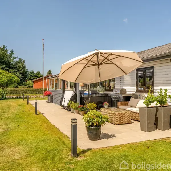 Parasol dækker terrassemøbler i en velplejet have ved en hvidmalet bolig. Solsenge og planter i krukker står rundt om bordet. Huset har store vinduer og har blå himmel.