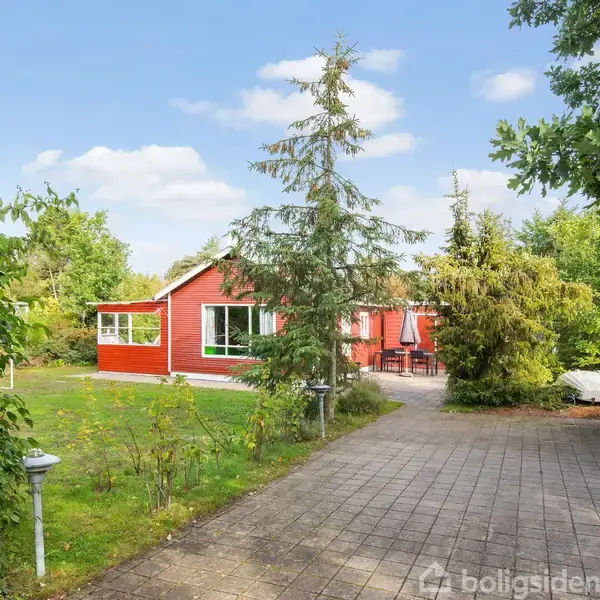 Et rødt træhus står stille i en frodig have med en flisebelagt sti i forgrunden. Omkring huset er der grønne buske og træer, der skaber en rolig atmosfære.