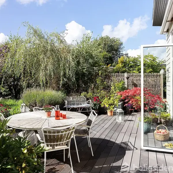 Rundt bord med stole står på en træterrasse, omgivet af frodig beplantning og farverige blomster. En åben glasdør forbinder terrassen med huset, mens solen skinner fra en blå himmel.