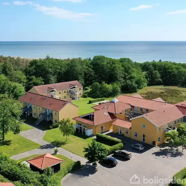 Flere gule bygninger med røde tage ligger i grønne omgivelser nær kysten. Baggrunden viser havet og en skov. Parkeringspladser er synlige foran bygningerne.