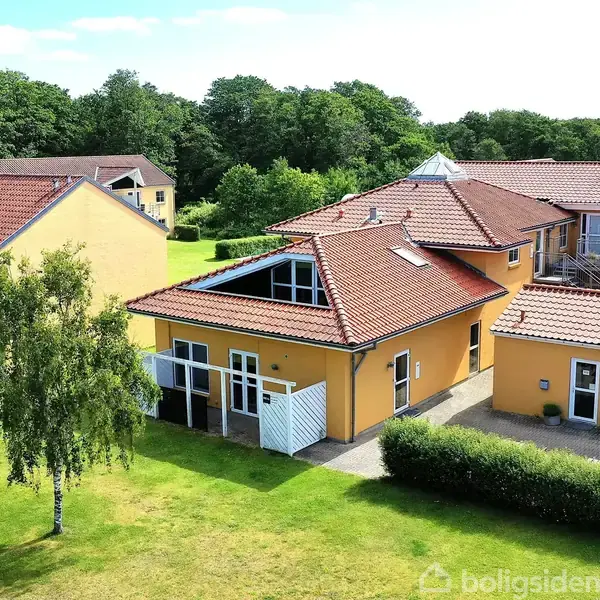 En række gule bygninger med røde tegltage ligger i et grønt område med træer og græs. Matcher de omkringliggende bygninger.