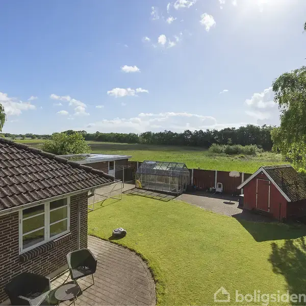 Hus af mursten med solpaneler på taget, beliggende i en grøn have med terrassemøbler. Baggrund med åbent landskab og små huse. Skarp sol og blå himmel uden skyer.