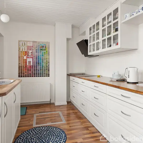 Et lyst køkken med hvide skabe og træbordplader. En elkedel og kaffemaskine står på bordet. På væggen hænger et farverigt maleri. Et rundt tæppe ligger på trægulvet.